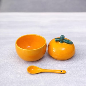 Può includere: Un set di condimenti in ceramica arancione, composto da una piccola ciotola, un contenitore con coperchio a forma di arancia con un gambo verde e un cucchiaino. Il set è progettato per servire salse o spezie.
