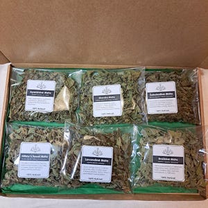 May include: A box containing six sealed bags of dried herbs, each with a white label. The labels feature text in a foreign language, along with the words "100% Natural." The herbs are green and appear to be mint varieties.