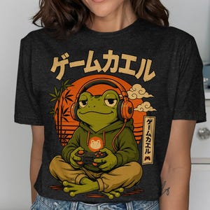 Op de afbeelding: Een antracietgrijs t-shirt met een afbeelding van een groene kikker die een oranje koptelefoon en een groene hoodie draagt terwijl deze een videogame speelt. Het ontwerp bevat een grote oranje zon, bamboestengels, wolken en Japanse tekens.