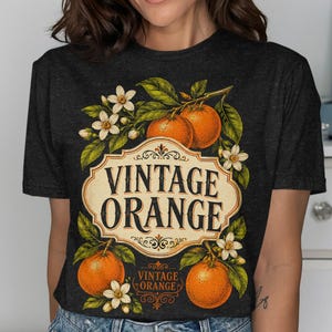 Puede incluir: Una camiseta gris carbón oscuro con un gráfico central de naranjas en ramas con flores blancas. El diseño incluye un marco de etiqueta decorativo de color crema con el texto VINTAGE ORANGE en una fuente serif negrita y texto más pequeño debajo.