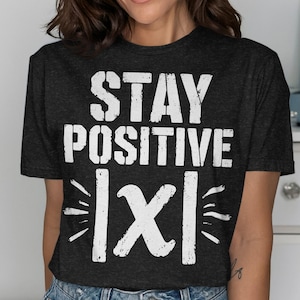 May include: A dark charcoal gray short-sleeve t-shirt featuring the white distressed text "STAY POSITIVE" above a graphic of the symbol "|X|" with small radiating lines on either side, printed in a bold, weathered, and textured white font.