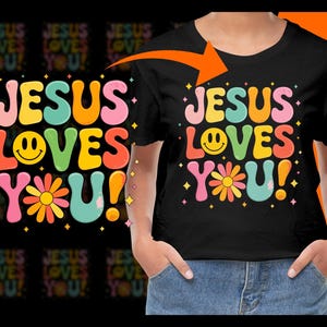 Può includere: Una maglietta nera con un design grafico colorato che dice "Jesus Loves You!" in un font retrò con una faccina sorridente e un fiore.