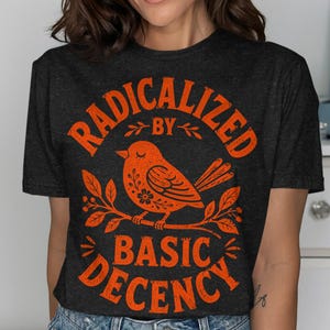 Può includere: Una t-shirt a maniche corte grigio antracite scuro con una grafica arancione vivace di un uccello appollaiato su un ramo. Il testo "RADICALIZED BY BASIC DECENCY" è stampato in un carattere arancione audace e usurato attorno all'uccello.