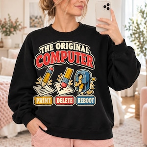 Puede incluir: Una sudadera negra de cuello redondo con el texto THE ORIGINAL COMPUTER. Debajo, las ilustraciones muestran un lápiz escribiendo con PRINT, una goma de borrar sobre papel con DELETE y un sacapuntas con la etiqueta REBOOT.