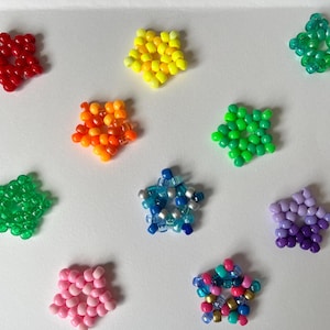 Puede incluir: Una variedad de cuentas en forma de estrella de colores, incluyendo rojo, amarillo, verde, naranja, azul, rosa y morado. Las cuentas están dispuestas sobre una superficie blanca, mostrando sus vibrantes colores y formas únicas.