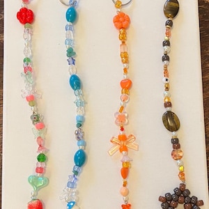 May include: Four beaded keychains with silver rings. The keychains feature various colourful beads, including red strawberries, blue mushrooms, orange flowers, and brown star shapes. The beads are strung together with clear string.