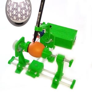 May include: A green 3D printed egg decorating machine with a brown egg in the centre. A black marker is held by a robotic arm above the egg. A white egg with a black geometric pattern is shown in the top left corner.
