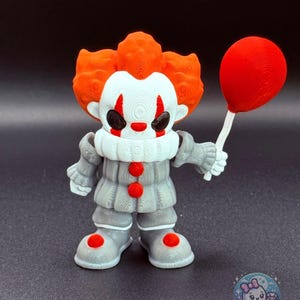 May include: A 3D-printed figurine of a clown with orange hair, white face, and gray outfit, holding a red balloon. The clown has red accents on the nose, cheeks, and shoes. The figurine is on a dark surface.