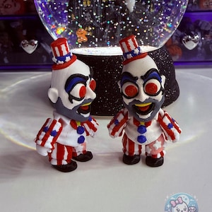 May include: Two small, clown-like figurines with red, white, and blue striped outfits and top hats. The figurines have exaggerated facial features, including red eyes and a painted beard. A snow globe with a Halloween theme is in the background.