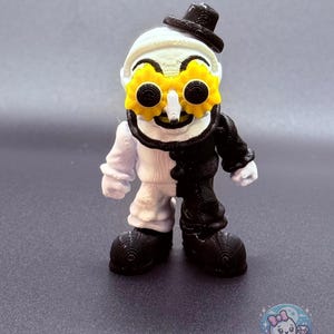 May include: A 3D-printed figurine of a whimsical character, split vertically with white and black halves. It wears a black top hat and yellow gear-shaped glasses. The figure has a white face, a black mustache, and large black shoes.