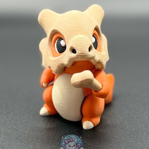 May include: A 3D-printed figurine of a Cubone character, featuring a bone helmet and orange body. The figurine has a cream-colored belly and is posed in a sitting position. The figurine is from BB's Bootique.