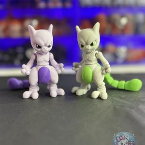 May include: Two 3D-printed figurines of a Pokemon character, one lavender and one light green. The lavender figure has a purple tail, and the green figure has a green tail. Both figures have a stern expression and are posed on a dark surface.