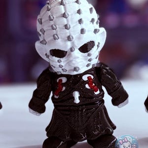 May include: A figurine of a character with a white face covered in gray pins, black eyes, and a black outfit with red accents. The figure has a detailed texture and is posed upright. The figurine is a collectible item.