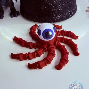 May include: A red, tentacled creature with a large, white and blue eyeball. The creature has a round head and eight ruffled tentacles. The background includes a black glittery object and a cartoon ghost logo.