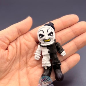 May include: A small, 3D-printed figurine of a clown with a black and white outfit. The clown has a white face with black eye makeup and a yellow smile. The figurine is held in a hand, with the text "BB's Bootique" visible.