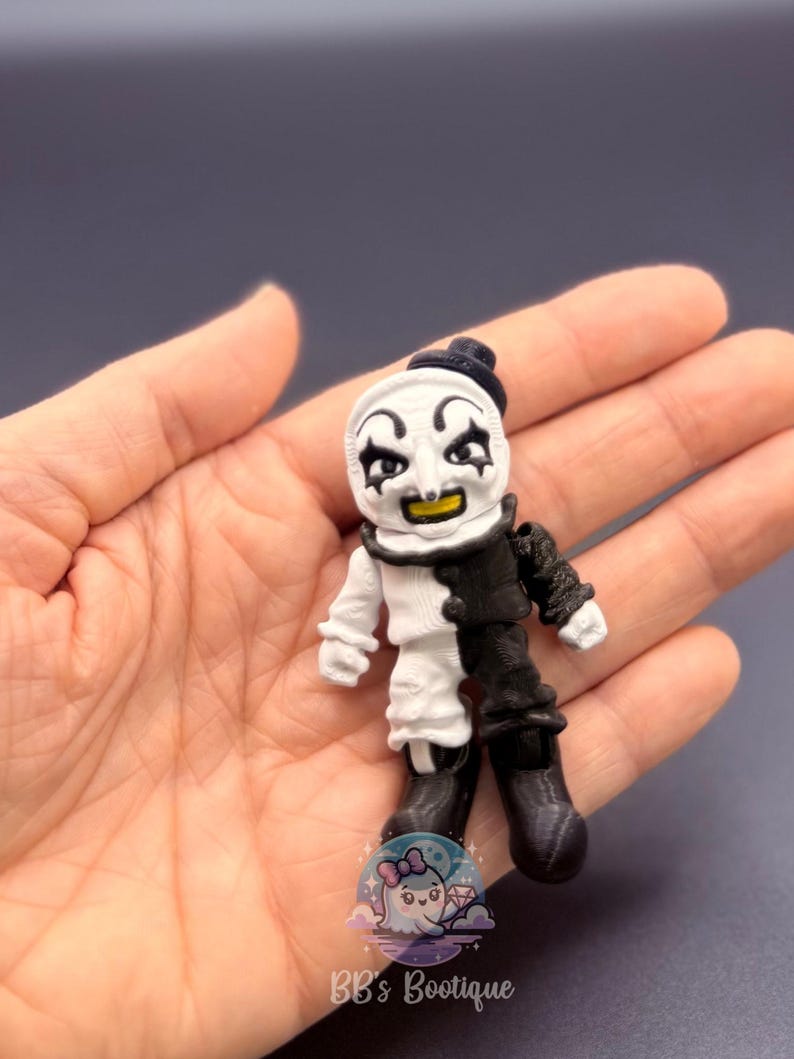 May include: A small, 3D-printed figurine of a clown with a black and white outfit. The clown has a white face with black eye makeup and a yellow smile. The figurine is held in a hand, with the text "BB's Bootique" visible.