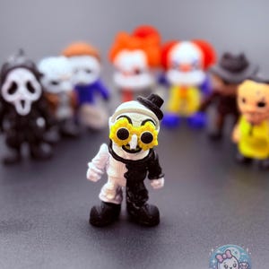 May include: A collection of miniature figurines featuring horror movie characters. The central figure is a clown-like character with a black and white split design, wearing a small black hat and yellow flower-shaped glasses. Other figures include a Ghostface, Pennywise, and other horror icons.