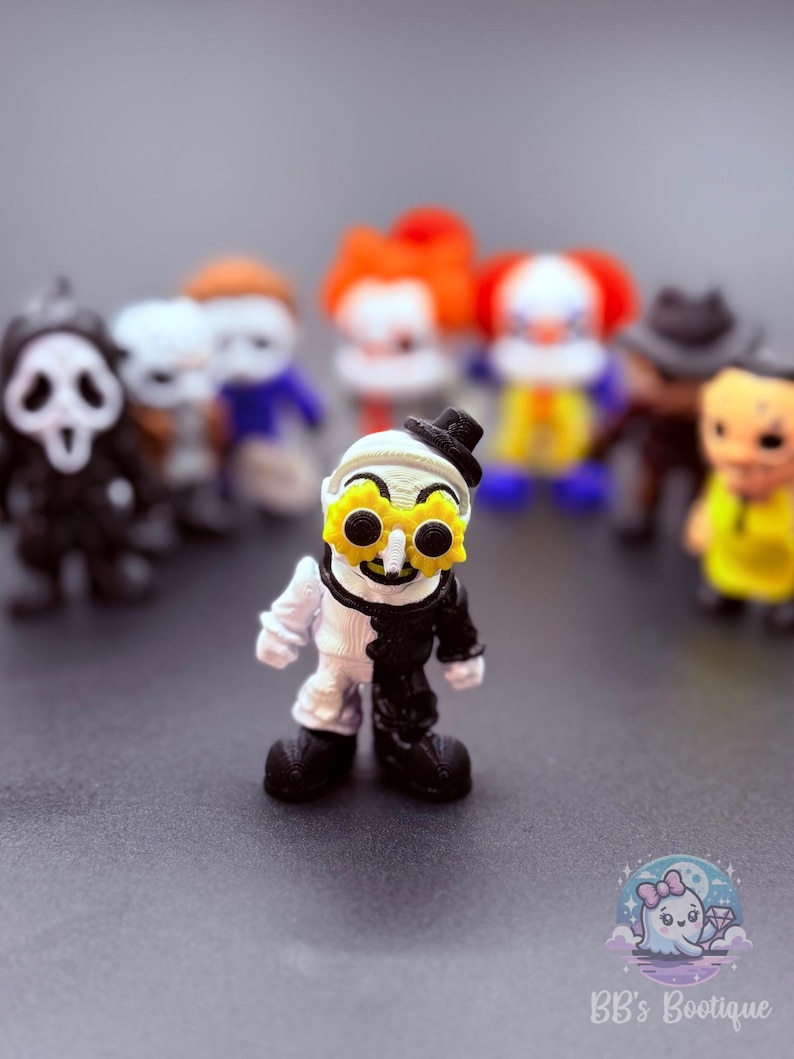 May include: A collection of miniature figurines featuring horror movie characters. The central figure is a clown-like character with a black and white split design, wearing a small black hat and yellow flower-shaped glasses. Other figures include a Ghostface, Pennywise, and other horror icons.