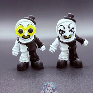 May include: Two small, detailed figurines of a clown with white skin, black and white clothing, and a small black hat. One clown wears yellow flower-shaped glasses. The figurines are on a gray surface.