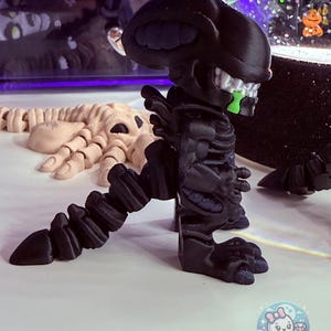 May include: A black alien figurine with a detailed, textured design. The alien has a large head, a skeletal face with green accents, and a segmented body. The figurine is posed on a white surface, with a light-colored skeleton figure in the background.