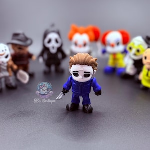 May include: A collection of miniature horror movie character figurines. The central figure is Michael Myers, holding a knife, in a blue jumpsuit. Other characters include Pennywise, Ghostface, and Freddy Krueger. The figures are set against a blurred gray background.