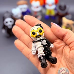 May include: A small, hand-held figurine of a clown with a black and white outfit and a yellow sunflower-shaped mask. Other figurines are blurred in the background. The figurine is held in a person's hand. The BB's Bootique logo is in the bottom right corner.