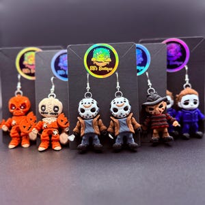 May include: A selection of Halloween-themed drop earrings showcasing miniature figures of horror movie characters. The earrings include characters such as Jason Voorhees, Freddy Krueger, and Michael Myers, set against a black backdrop.
