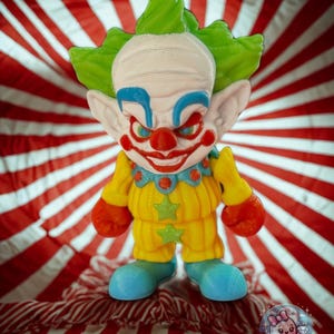 May include: A colorful clown figurine with a white head, green hair, and a red nose. The clown wears a yellow outfit with blue accents and orange hands. The background is a red and white striped pattern.