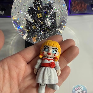 May include: A Halloween-themed snow globe with a black haunted house and white ghosts inside. A small doll with blonde hair, blue eyes, and a white dress is in front of the snow globe. The doll has a red ribbon and a red rose.
