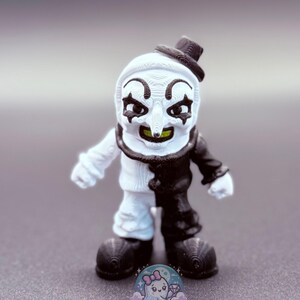 May include: A small figurine of a clown with a white and black color scheme. The clown has a white face with black eye markings, a long nose, and a small black hat. The clown's outfit is split, with one side white and the other black.