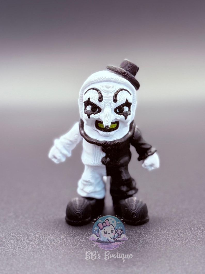 May include: A small figurine of a clown with a white and black color scheme. The clown has a white face with black eye markings, a long nose, and a small black hat. The clown's outfit is split, with one side white and the other black.