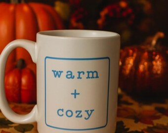 May include: White ceramic mug with the words "warm + cozy" in blue, set against a backdrop of pumpkins and fall foliage. The mug has a large handle and a square design with the text.