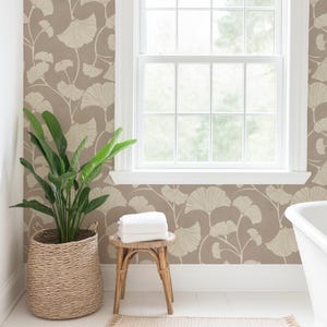 May include: Wallpaper featuring a repeating pattern of cream-colored ginkgo leaves on a muted taupe background. The design covers the wall surrounding a white-framed window with a bamboo shade, positioned above a woven basket with a green plant.