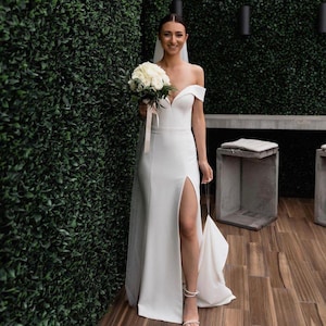 Puede incluir: Un vestido de novia blanco de hombros descubiertos con una abertura en el muslo y una cola larga. El vestido está hecho de una tela lisa y satinada. La novia lleva un ramo de flores blancas.