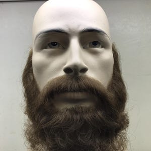 May include: A mannequin head with a bald scalp, realistic facial features, and a full, brown beard. The beard is styled with a thick mustache and sideburns, and it has a natural, textured appearance. The mannequin is set against a neutral background.