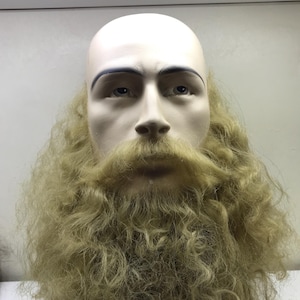 May include: A mannequin head with a bald scalp, blue eyebrows, and a long, curly, light-colored beard and mustache. The mannequin has a neutral expression and is set against a plain background.