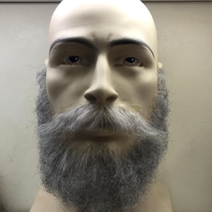 May include: A mannequin head with a full, gray beard and mustache. The beard is thick and covers the chin and jawline. The mannequin has a neutral expression and is set against a plain background.