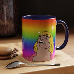 May include: A ceramic mug with a dark blue interior and handle, featuring a rainbow gradient background and a cartoon illustration of a blonde character with a pink shirt. The design includes white four-pointed star accents scattered around the figure.