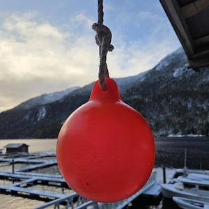 May include: A bright red buoy hangs from a rope against a backdrop of a snowy mountain range and a calm body of water. The buoy is round with a bulbous shape and is suspended from a knotted rope. The scene suggests a coastal or marine environment.