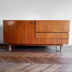 Puede incluir: Un aparador rectangular de madera con un acabado marrón claro y vetas de madera verticales. La unidad incluye una puerta de armario grande a la izquierda con un pequeño pomo metálico y dos cajones horizontales apilados en el lado derecho.