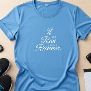 May include: A light blue athletic t-shirt with the text "If you Run you're a Runner" in white script. The shirt is on a light wood surface, with running shoes, a watch, and a water bottle nearby.