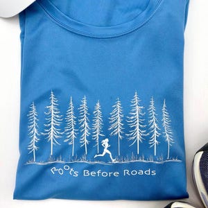 May include: A blue athletic shirt with a white graphic of a runner and pine trees. The text "Roots Before Roads" is printed below the graphic. The shirt is folded and appears to be made of a moisture-wicking material.