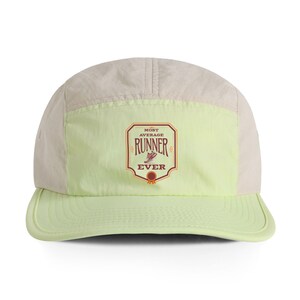 May include: A five-panel cap in cream and light green. The front panel features a rectangular emblem with the words "MOST AVERAGE RUNNER EVER" and a running shoe graphic in brown and red.