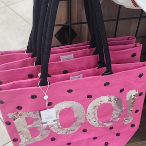 May include: A bright pink tote bag featuring black polka dots and the word BOO! in large silver sequins. The bag has two black fabric handles and a white product tag attached to one handle. The interior features a small white brand label.