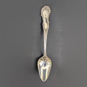 May include: A silver spoon with an ornate handle and a rounded bowl. The handle features a decorative floral design and a small, engraved area. The spoon is set against a dark gray background, highlighting its polished surface.