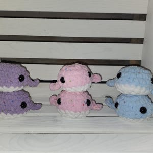 May include: Three pairs of crocheted whale plushies. One pair is purple and white, one pair is pink and white, and one pair is blue and white.