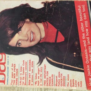 Può includere: Una copertina di rivista con una donna con lunghi capelli scuri che indossa un maglione rosso a collo alto e una giacca nera. La rivista si intitola "Woman's Day" e ha il testo "How Jackie Onassis-and the other beautiful people-stay that way (and how you can too)" sul lato destro della copertina.