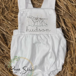 Puede incluir: Romper blanco para bebé con tirantes y parte inferior redondeada. La parte delantera presenta un diseño de perro de caza bordado con el nombre "hudson" en escritura cursiva. El romper tiene cierres de botón y está hecho de tela suave.