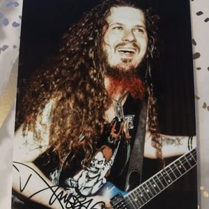 May include: A black and white photo of a musician with long, curly hair playing a blue electric guitar. The musician is wearing a black shirt with a skull graphic and a signature that reads "Dimebag".