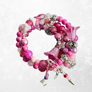Peut inclure: Un bracelet à plusieurs rangs avec un assortiment de perles roses, rondes, facettées et texturées. Le bracelet est orné de breloques en forme de fleurs, d'éléments argentés et de décorations pendantes. La palette de couleurs comprend des nuances de rose, de blanc et d'argent.